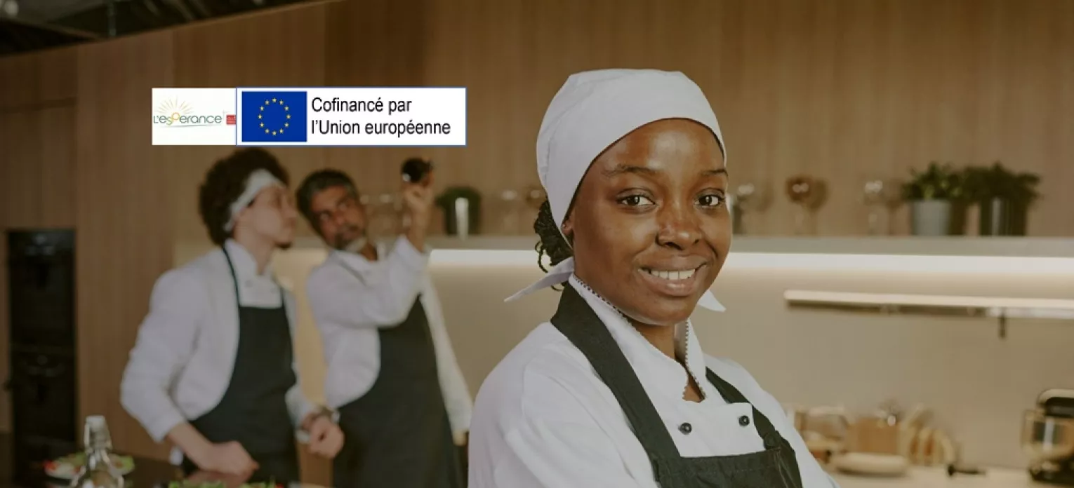 Jeune fille en tenue de cuisinière au 1e plan dans une cuisine. Deux autres personnes sont présentes en arrière-plan, également en tenue. 